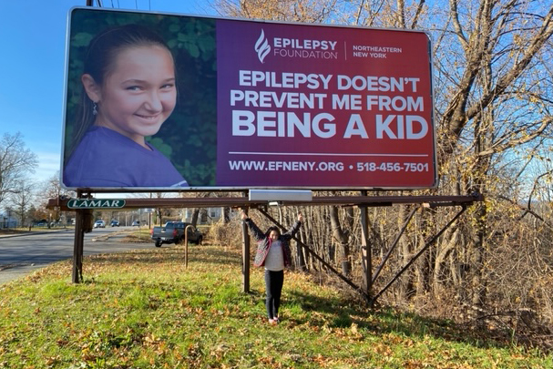 Green Meadow 5th grader Madison Pospisil standing in front of a billboard featuring her as a 2023 Winning Kid for the Epilepsy Foundation of Northeastern New York.